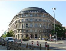 Großes, halbrundes Gebäude aus Stein mit mehreren Stockwerken und einer dunkel gedeckten Kuppel. Das Gebäude befindet sich an einer Straßenecke, vor der Verkehrszeichen, Absperrungen und ein Gehweg mit Autos, Fahrrädern und Fußgängern zu sehen sind.