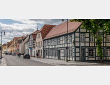 Straßenszene mit einer Reihe historischer Fachwerk- und Putzgebäude, die eng aneinander stehen. Die Fassaden sind in hellen Farben gehalten, mit Ziegeldächern und teilweise geschmückten Fenstern; links parken Autos, während rechts eine Baumreihe den Gehweg säumt.