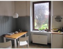 Kleine Küche mit Holztisch und zwei weißen Stühlen, daneben ein großes Fenster mit Blick auf einen Garten. Die Wände sind teilweise mit grauem Holz vertäfelt, es gibt eine Hängelampe, eine Heizung unter dem Fenster und eine Arbeitsfläche mit Obst und Küchenmessern an der Wand.