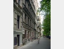 Straßenszene mit Altbauten auf der linken Seite, teilweise mit sichtbaren Schäden an der Fassade, und Reihen von Balkonen. Rechts des Gehwegs stehen Bäume, die Schatten spenden, und vereinzelte Passanten sowie geparkte Fahrräder sind zu sehen.