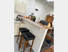 Eine moderne Küche mit einer erhöhten Holztheke, an der zwei gepolsterte Barhocker mit schwarzem Sitz stehen. Auf der Theke befinden sich ein Brotkasten, Messerblöcke, eine Zimmerpflanze und verschiedene Lebensmittel. Im Hintergrund sind weiße Wände, ein Holzschrank und ein metallisches Treppengeländer sichtbar.