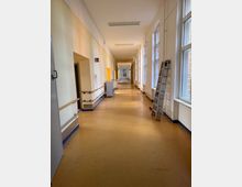 Langer, schmaler Flur mit gelbem Bodenbelag und hellen Wänden, links Handläufe sowie Türen, rechts hohe Fenster mit viel Tageslicht. Eine Anlegeleiter lehnt an der rechten Wand, und am Ende des Flurs ist eine Glastür sichtbar.