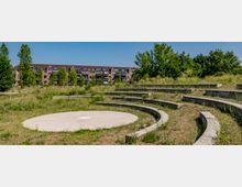 Eine halbrunde, terrassenförmige Freilichtbühne mit Sitzreihen aus Beton, umgeben von Wiesen und Bäumen im Vordergrund. Im Hintergrund sind mehrstöckige Wohngebäude mit Balkonen zu sehen.