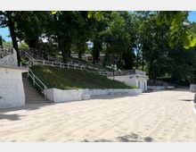Sandy open area bordered by low white retaining walls and a sloped hillside with stairs and white railings. Dense trees line the upper terrace, with small white utility buildings and lampposts visible along the path.