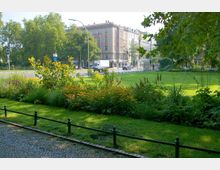 Städtischer Park mit gepflegtem Rasen, Blumenbeeten und Bäumen im Vordergrund; im Hintergrund eine Straße mit Autos, Fahrradständern und mehrstöckigen Wohngebäuden. Links sind ein Gehweg und eine Bushaltestelle sichtbar.