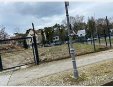 Freifläche hinter einem schwarzen Metallzaun, umgeben von Gehwegen mit Pflastersteinen. Im Hintergrund stehen Bäume und Wohnhäuser, während parkende Autos an einer Straße sichtbar sind. Der Himmel ist bewölkt.