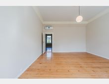 Leerer, heller Wohnraum mit hohen Decken, weißen Wänden und breitem Holzdielenboden. Eine runde Deckenleuchte hängt mittig, und durch eine offene Tür ist ein angrenzender Raum mit Fenster zu sehen.