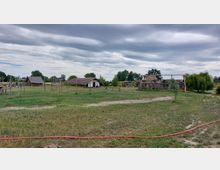 Freilichtmuseum mit historischen Holzbauten und strohgedeckten Hütten auf einer großen Wiese, umgeben von Bäumen und weiten Feldern unter einem bewölkten Himmel. Im Vordergrund befinden sich Pfähle und ein schmaler Pfad.