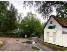 Ein ländlicher Weg mit Pfützen führt an einem weißen Gebäude mit der Aufschrift „Gartenlokal-Vereinshaus Mühlenwiese-Finkenherd“ vorbei. Im Hintergrund stehen ein kleines Haus mit Spitzdach und Bäume, die von dichtem Grün umgeben sind.