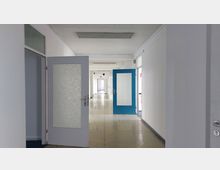 Langer, schlichter Innenflur eines Gebäudes mit weißen Wänden, offenstehenden Türen und einfachem Linoleumboden. Im mittleren Bereich befindet sich eine blaue Tür mit Glaseinsatz, während die Beleuchtung durch Deckenleuchten gewährleistet wird.