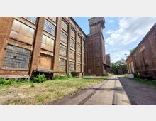 Ehemaliges Fabrikgelände mit roten Backsteingebäuden und großen, teilweise beschädigten Fenstern. Im Vordergrund verläuft ein Weg mit Gleisen, umgeben von vereinzelter Vegetation.