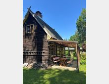 Holzhaus mit steilem Satteldach und kleinem Dachfenster, daneben eine überdachte Veranda mit Sitzbank und Tisch. Das Gebäude steht in einem grünen Garten mit Rasen und Bäumen unter klarem blauem Himmel.