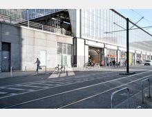 Straßenansicht eines modernen Bahnhofsgebäudes mit einer Glasfassade und metallischen Strukturelementen. Vor dem Gebäude befinden sich Gehwege, Fahrradständer mit abgestellten Fahrrädern und Straßenbahnschienen im Vordergrund.
