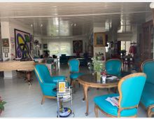 Ein heller Innenraum mit einer Mischung aus Wohn- und Kunstgalerie-Atmosphäre, ausgestattet mit türkisfarbenen Polsterstühlen und einem runden Holztisch. An den Wänden hängen farbenfrohe Gemälde, und im Hintergrund sind große Fenster mit Blick auf eine Gartenlandschaft sichtbar.