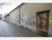 Gepflasterte Straße entlang einer langen, schlichten Mauer mit Graffiti und einer Holztür, umgeben von einer urbanen Umgebung. Im Hintergrund sind Wohngebäude und ein geparktes Auto sichtbar.