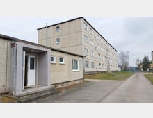 Mehrstöckiges Wohngebäude aus Betonplatten mit rechteckigen Fenstern und einer kleinen überdachten Eingangsverlängerung mit einer weißen Tür. Das Umfeld besteht aus einem gepflasterten Weg, Rasenflächen und einigen Bäumen im Hintergrund.