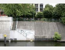 Uferbereich mit einer betonierten Böschung, die von einer Treppe und einem Geländer durchbrochen wird, versehen mit Graffiti. Im Vordergrund fließt ein Kanal, am Wasser steht ein gelbes Geländer, und im Hintergrund sind Bäume sowie ein mehrstöckiges Wohngebäude sichtbar.