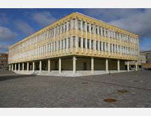 Eckiges, zweistöckiges Gebäude im Stil der Moderne mit gelber Fassade und vertikalen Betonstreben vor den Fenstern; es steht auf weißen Säulen und ist von einem gepflasterten Platz umgeben.