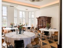 Heller Bankettsaal mit großen Sprossenfenstern und Stuckdecke, ausgestattet mit runden Tischen mit weißen Tischdecken, Gedecken und Blumenvasen. An der Wand steht ein großer dunkler Holzschrank; der Boden ist ein Parkett im geometrischen Muster.