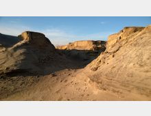 Wüstenähnliche Landschaft mit sandigen, erodierten Felsformationen und einer von Schatten durchzogenen Senke. Im Hintergrund ist ein klarer blauer Himmel zu sehen.