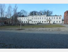 Breiter, gepflasterter Platz mit einem weißen zweigeschossigen Gebäude im Hintergrund, das große Fenster und zwei Balkone aufweist. Im Vordergrund stehen vereinzelt junge Bäume, sowie eine Rasenfläche, und dahinter befinden sich Bäume und ein Backsteingebäude am rechten Bildrand.