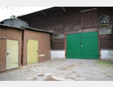 Außenansicht eines landwirtschaftlichen Gebäudes mit brauner Holzverkleidung, einem großen grünen Tor und zwei gelben Türen an einer angrenzenden Wand. Im Hintergrund ist ein metallener Wassertank sichtbar.