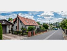 Einfamilienhäuser mit roten Ziegeldächern und Vorgärten entlang einer ruhigen, asphaltierten Straße, gesäumt von Bäumen und Zäunen; links im Vordergrund eine Garage mit einem Wegweiser „Schmiedeweg“.