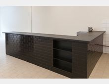 Moderne Küchen- oder Barzeile in L-Form mit schwarzer Arbeitsplatte und Fronten aus glänzenden, schwarzen Rechteckfliesen, inklusive offenem Regalfach. Heller Raum mit beigem Fliesenboden und weißen Wänden, im Hintergrund teils weiß geflieste Wandbereiche.