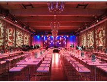 Großer Veranstaltungsraum mit einer langen Tischreihe in der Mitte und hohen Barhockern, dekoriert mit Gläsern, Flaschen und roten Blumen. Die Backsteinwände sind mit Lichtprojektionen verziert, und im Hintergrund ist eine beleuchtete Bühne zu sehen.