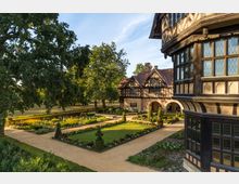 Historisches Gebäude im Tudorstil mit Fachwerk und Arkaden, umgeben von einem gepflegten Garten mit symmetrischen Wegen, Blumenbeeten und Hecken. Im Hintergrund stehen große Bäume, die das Anwesen einrahmen.
