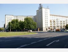 Wohn- und Geschäftsgebäude im Stil des Sozialistischen Klassizismus, umgeben von Bäumen am Straßenrand, mit einer mehrspurigen Straße und Kreisverkehr im Vordergrund. Die Fassade des Gebäudes ist mit großen Fenstern, Balkonen und rechtwinkligen Linien gestaltet.