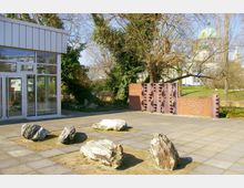 Außenbereich mit gepflastertem Boden, vereinzelten großen Steinen als Dekoration und einem modernen Gebäude mit großen Glasfenstern auf der linken Seite. Im Hintergrund stehen Bäume, ein gemauerter Zaun aus roten Ziegeln und eine Kuppel eines historischen Gebäudes, teilweise verdeckt durch die Vegetation.