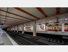Bahnhofsplattform einer U-Bahn-Station mit zwei Gleisen und mittigem Bahnsteig, umgeben von hell gekachelten Wänden und orangeroten Deckenstreben. Eine S-Bahn fährt gerade ein, und einige Passanten sind mit Gepäck auf dem Bahnsteig zu sehen.