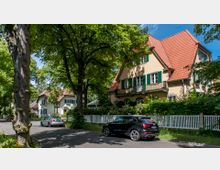 Wohnstraße mit großen, freistehenden Häusern im traditionellen Stil, gekennzeichnet durch rote Ziegeldächer, grüne Fensterläden und weiße Zäune. Die Straße ist gesäumt von hohen Bäumen, und geparkte Autos stehen entlang der Fahrbahn.