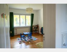 Blick durch eine offene Tür in ein helles Kinderzimmer mit Holzfußboden, großer Fensterfront und grünen Vorhängen. In der Mitte stehen ein kleiner blauer Kindertisch mit zwei Stühlen auf einem bunten Teppich; rechts befindet sich ein niedriges Bücherregal.