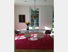 Esszimmer mit ovalem weißen Marmortisch, umgeben von weißen Stühlen mit Holzbeinen, auf einem pinken Teppich. Im Hintergrund stehen zwei hohe Fenster mit Blick ins Grüne, darüber hängt ein Kronleuchter, und links ist ein Gemälde an der weißen Wand zu sehen.