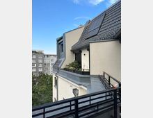 Blick von einem Balkon auf das Dachgeschoss eines hell verputzten Wohnhauses mit steilem Ziegeldach, Dachfenstern und Regenrinne sowie einer kleinen bepflanzten Dachterrasse mit schwarzem Geländer. Im Hintergrund sind ein mehrstöckiges Wohngebäude, Baumwipfel und ein blauer Himmel zu sehen.