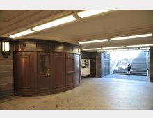 Unterirdischer Durchgang mit braunem Holzpavillon und Treppenaufgang, der ins Freie führt. Beleuchtung durch lineare Deckenleuchten, links eine Wandleuchte und rechts ein schmiedeeisernes Tor.