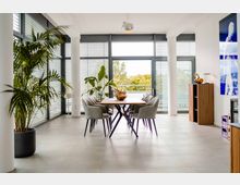 Großzügiger, moderner Essbereich mit einem Holztisch und gepolsterten Stühlen, umgeben von bodentiefen Fenstern mit Jalousien. Im Raum stehen Pflanzen und ein Holzschrank, während durch die Fenster eine Terrasse mit Blick ins Grüne sichtbar ist.