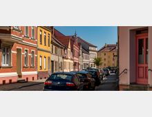 Straßenszene in einem Wohngebiet mit Häusern in verschiedenen Farben wie Rot, Gelb und Beige, teils mit Backsteinfassaden und roten Ziegeldächern. Geparkte Autos säumen die Pflasterstraße, und im Hintergrund ist ein kleiner Baum zu sehen.