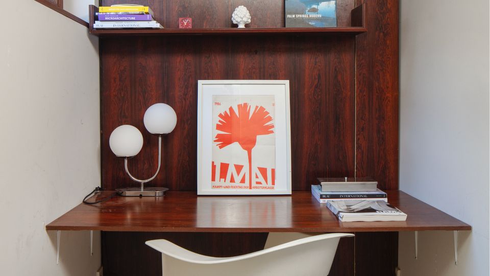 Small built-in home office nook with a dark wood desk and wall-mounted shelving, set between white walls under an exposed concrete ceiling. A window on the left brings in daylight, and the desk holds a globe lamp, stacked books, and framed artwork, with a white pedestal chair in front.
