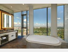 Modernes Badezimmer mit freistehender Badewanne vor einer großen Fensterfront, die einen Panoramablick auf eine bewaldete Landschaft und die Stadt bietet. Im Vordergrund ein Waschbereich mit beleuchtetem Spiegel und Marmordetails.