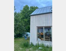 Außenansicht eines hellen Holzhauses mit steilem Metalldach und großem Fenster zur Gartenseite. Daneben liegt eine Wiese mit Sitzmöbeln, im Hintergrund stehen hohe Bäume und ein kleines Gewächshaus.