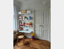 Ein heller Raum mit einer weißen Wand, an der offene Regale mit Büchern, Bastelmaterialien und Aufbewahrungsboxen angebracht sind. Rechts befinden sich weiße Doppeltüren mit verzierter Paneelgestaltung, und der Boden besteht aus herringbone-verlegtem Holzparkett. Ein Holzstuhl steht vor einem Regaltisch, daneben bunte Spielscheiben aufgestapelt.