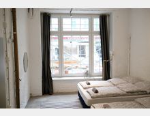 Ein schlicht eingerichtetes Zimmer mit weiß gestrichenen Wänden, einem großen Fenster mit Sprossen, das von schwarzen Vorhängen eingerahmt wird. Im Vordergrund stehen zwei nebeneinanderliegende Betten mit grauer Bettwäsche, und durch das Fenster sind Baustellenleitplanken sichtbar.