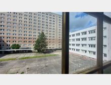 Blick auf einen rechteckigen Innenhof mit Betonboden, umgeben von einem mehrstöckigen, beigen Wohngebäude links und einem weißen Verwaltungsgebäude rechts. Im Innenhof stehen ein Baum, Grünflächen und ein Zaun im Vordergrund.