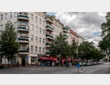 Mehrstöckiges Wohngebäude mit heller Fassade und grünen Balkongeländern, die teilweise mit Pflanzen dekoriert sind. Im Erdgeschoss befinden sich Ladenlokale mit roten Markisen; ein Gehweg mit Gehölz und parkenden Autos grenzt an die Straße.