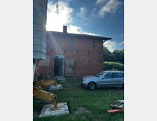 Backstein-Gebäude mit zwei Etagen, teilweise renovierungsbedürftig, auf einem grasbewachsenen Grundstück. Im Vordergrund stehen ein silbernes Auto und Teile eines Baugeräts, während die Umgebung von Bäumen umgeben ist.