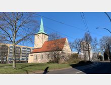 Historische Kirche mit Feld- und Ziegelsteinmauern sowie einem roten Satteldach, ergänzt durch einen hellen Turm mit grüner Spitze, umgeben von kahlen Bäumen auf einer Rasenfläche; im Hintergrund moderne Wohn- und Bürogebäude.