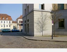 Straßenecke mit gepflastertem Boden, flankiert von modernen, hellen Gebäuden. Im Hintergrund sind weitere historische Gebäude mit roten Ziegeldächern sichtbar, geparkte Autos stehen entlang der Straße, und Straßenschilder sowie ein Laternenmast befinden sich im Vordergrund.